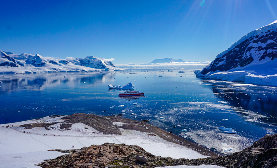 A gorgeous view from the top of a hill in Antarctica. You can see distant icy mountains, crystal royal blue waters and a red and white expedition ship in the water.