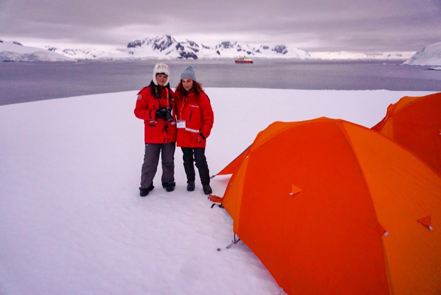 2 girls bundled up standing on a snowy island next to a tent in Antarctic with the ship in the background on the water.