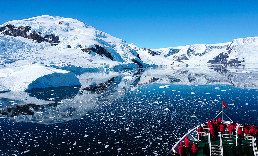 A Ship pulls into a harbor in Antarctica with the icy mountains refected in the water with chunks of ice floating