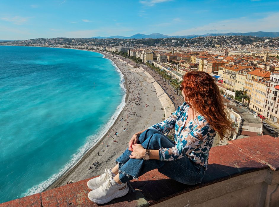Red head woman sits on ledge looking down at the seaside in Nice, France